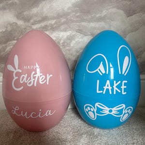 May include: Two large plastic Easter eggs, one pink and one blue. The pink egg has "Happy Easter" and "Lucia" in white script. The blue egg features bunny ears, a bow tie, paw prints, and the name "LAKE" in white.