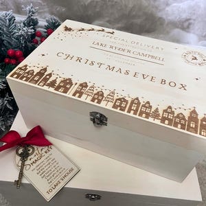 May include: A white wooden Christmas Eve box with engraved details. The box features a decorative village scene along the bottom edge and text that reads "CHRISTMAS EVE BOX". A tag with a key and red ribbon is attached. The box is designed for special delivery.