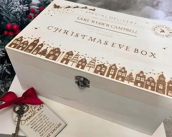 May include: A white wooden Christmas Eve box with engraved details. The box features a decorative village scene along the bottom edge and text that reads "CHRISTMAS EVE BOX". A tag with a key and red ribbon is attached. The box is designed for special delivery.