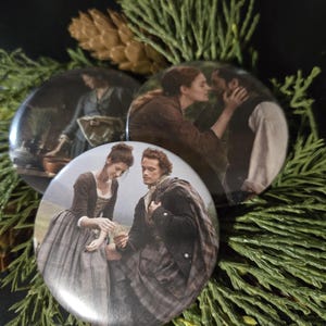 Peut inclure: Trois épingles rondes représentant différents couples de la série télévisée Outlander. La première épingle montre une femme en robe bleue avec un homme en veste marron. La deuxième épingle montre une femme en robe rouge avec un homme en veste noire. La troisième épingle montre une femme en robe blanche avec un homme en veste noire.