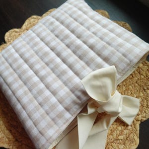 May include: A beige and white checked quilted pillow with a cream-coloured bow. The pillow is placed on a woven placemat with a scalloped edge. The pillow's fabric has a grid pattern.