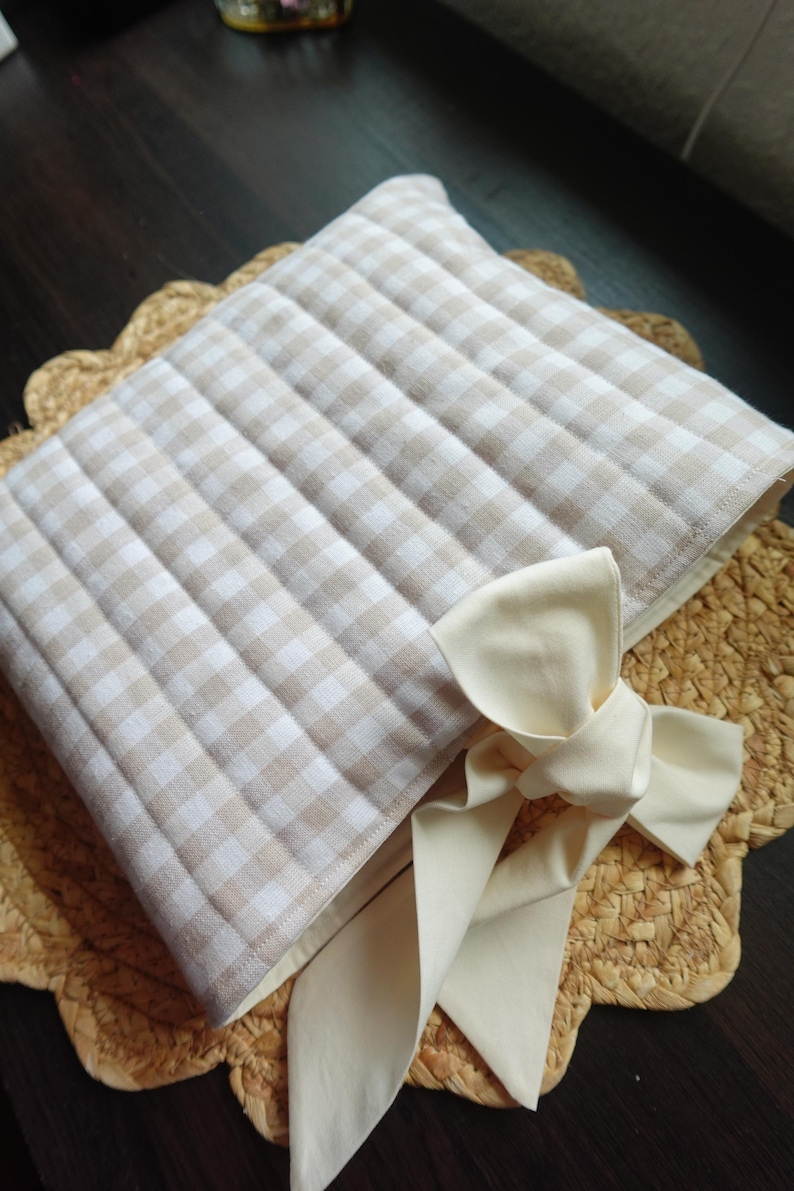 May include: A beige and white checked quilted pillow with a cream-coloured bow. The pillow is placed on a woven placemat with a scalloped edge. The pillow's fabric has a grid pattern.