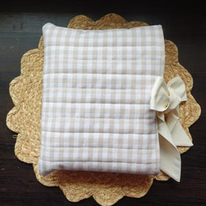 May include: A beige and white gingham patterned pillow with a cream-coloured bow, resting on a woven placemat with a scalloped edge. The pillow has horizontal stitching.