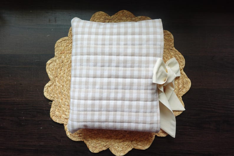 May include: A beige and white gingham patterned pillow with a cream-coloured bow, resting on a woven placemat with a scalloped edge. The pillow has horizontal stitching.