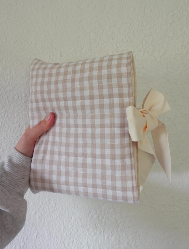 May include: A beige and white gingham patterned clutch with a cream-coloured bow. The clutch is rectangular and appears to be made of fabric. The bow is tied on the side of the clutch.