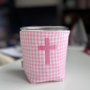May include: A glass jar with a pink and white gingham fabric sleeve. The sleeve features an embroidered pink cross. The jar sits on a white surface.