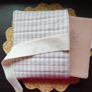 May include: A light pink leather journal with floral embossing, secured with a cream fabric tie, rests on a beige and white checked quilted cover. The journal and cover are placed on a woven placemat.