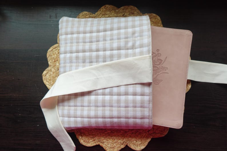 May include: A light pink leather journal with floral embossing, secured with a cream fabric tie, rests on a beige and white checked quilted cover. The journal and cover are placed on a woven placemat.