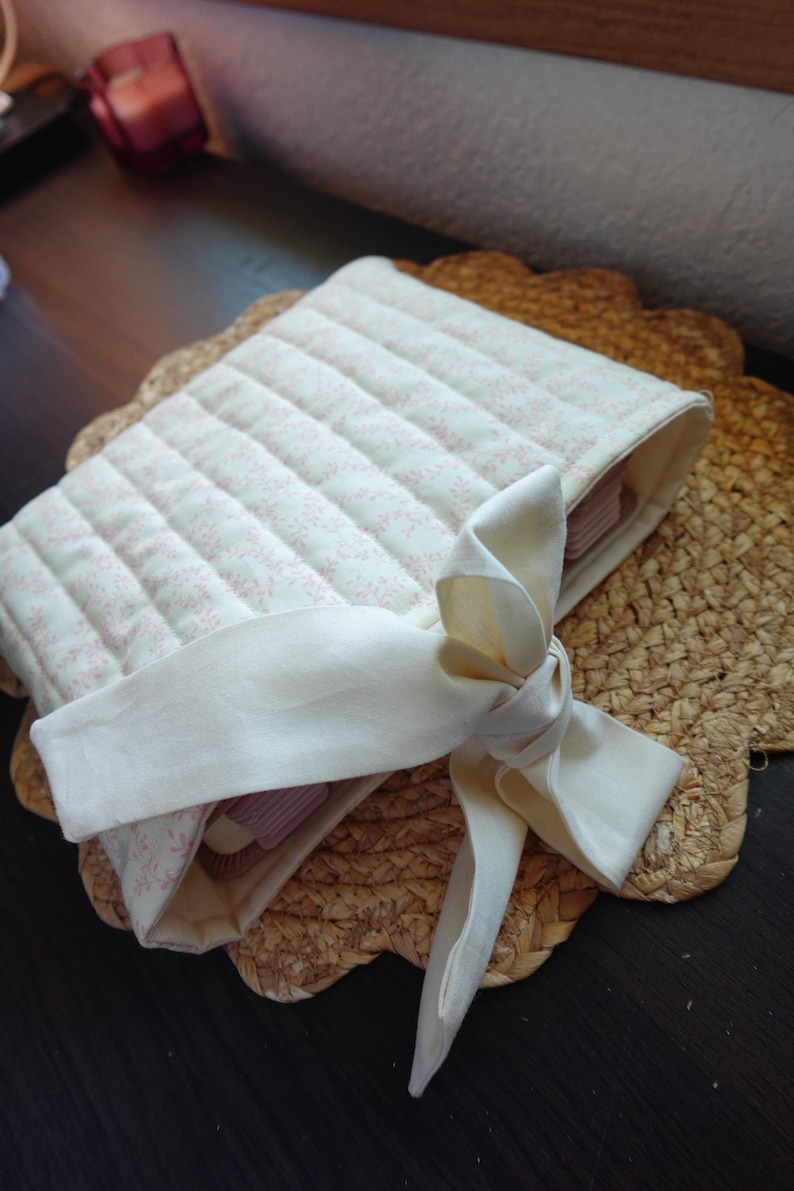 Op de afbeelding: Een witte, gestikte boekomslag van stof met een roze bloemenpatroon en een wit lint dat in een strik is gebonden. De omslag ligt op een geweven stro mat.