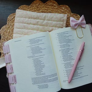 Op de afbeelding: Een open witte en roze Bijbel met roze tabs en een roze pen. Een roze strik wordt gebruikt als bladwijzer. De Bijbel ligt op een geweven strooien placemat.