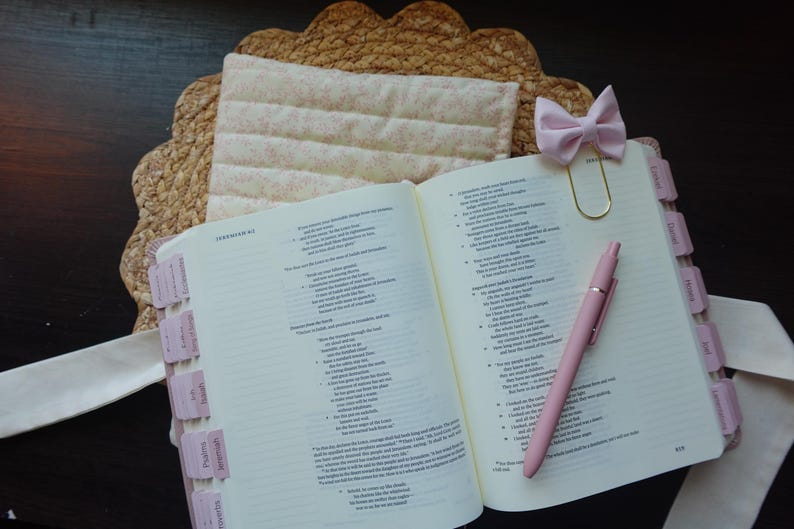 Op de afbeelding: Een open witte en roze Bijbel met roze tabs en een roze pen. Een roze strik wordt gebruikt als bladwijzer. De Bijbel ligt op een geweven strooien placemat.