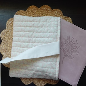 Op de afbeelding: Een roze en witte quilted boekomslag met een witte lint sluiting. De omslag is ontworpen om een ​​paars lederen dagboek met een reli&euml;f bloemenontwerp te houden.