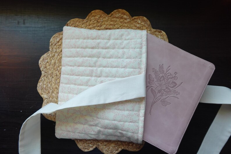 Op de afbeelding: Een roze en witte quilted boekomslag met een witte lint sluiting. De omslag is ontworpen om een ​​paars lederen dagboek met een reli&euml;f bloemenontwerp te houden.