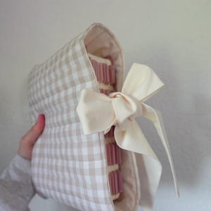 May include: A beige and white gingham fabric book cover with a cream-coloured bow. The cover is wrapped around a stack of pink-edged pages. The bow is tied on the side, adding a decorative touch to the book cover.
