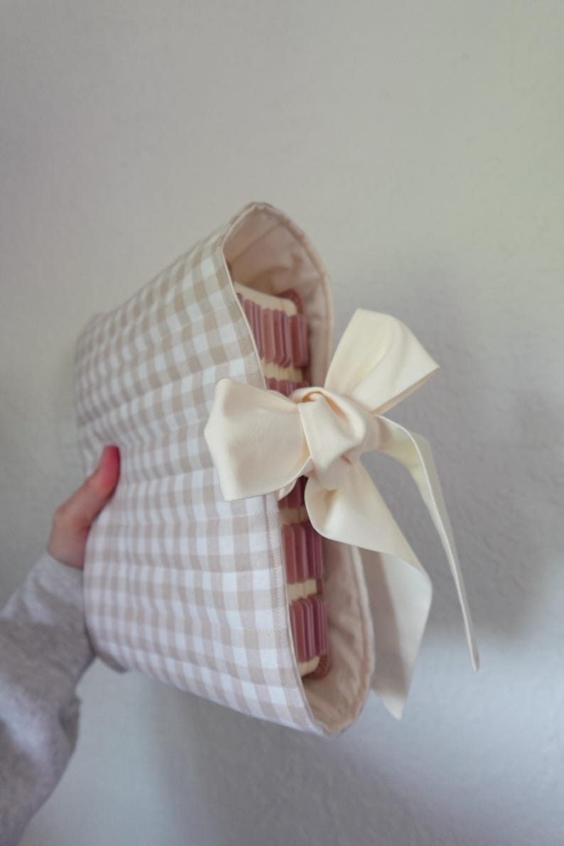 May include: A beige and white gingham fabric book cover with a cream-coloured bow. The cover is wrapped around a stack of pink-edged pages. The bow is tied on the side, adding a decorative touch to the book cover.