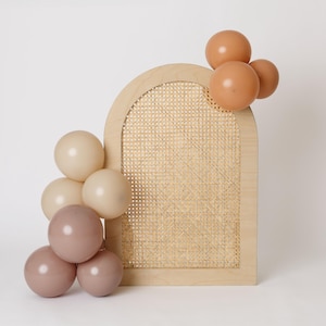 May include: A wooden archway with a woven cane center, adorned with clusters of balloons in shades of beige, tan, and mauve. The balloons are arranged around the arch, creating a decorative display. The arch is a light wood color.