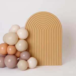 May include: A beige arch-shaped decorative piece with a series of parallel lines, next to a cluster of beige, peach, and mauve-colored spheres. The objects are set against a white background, creating a minimalist aesthetic.