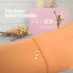 Könnte beinhalten: Ein goldfarbenes Armband mit einem Weinglas-Anhänger. Eine rosa Karte mit dem Text "Für deine beste Freundin" und "Holla die WEIN fee" befindet sich im Hintergrund. Die Karte zeigt auch eine goldene Illustration einer Fee.