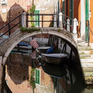 May include: A stone arch bridge over a canal in Venice, Italy. Two small boats are docked under the bridge. The reflection of the buildings and the bridge are visible in the water.
