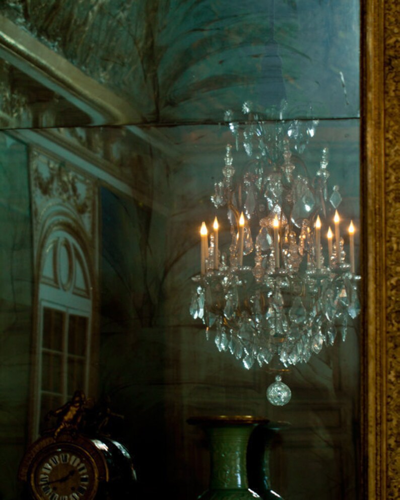 May include: A crystal chandelier with lit candles reflected in a mirror. The mirror is framed in gold and shows a room with ornate details.