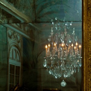 May include: A crystal chandelier with lit candles reflected in a mirror. The chandelier is hanging from the ceiling and is surrounded by a dark green background.