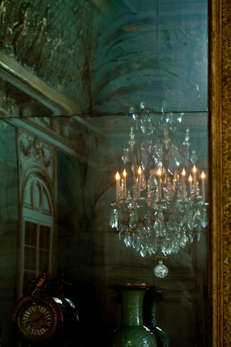 May include: A crystal chandelier with lit candles reflected in a mirror. The chandelier is hanging from the ceiling and is surrounded by a dark green background.
