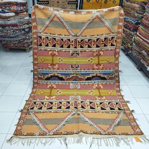 May include: A colorful Moroccan rug with geometric patterns in shades of orange, yellow, pink, blue, and black. The rug has a fringe border and is woven with wool.