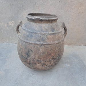 Peut inclure: Un grand récipient en terre cuite brun foncé, de forme arrondie, doté d'une large ouverture et de deux petites anses robustes sur les côtés. La surface présente un aspect patiné et texturé avec des tons bruns nuancés et des stries.