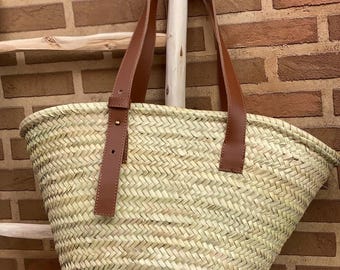 May include: Woven straw tote bag with brown leather handles and accents. The bag has a rectangular shape with a wide opening and is designed for carrying items. The bag is made of natural materials and has a rustic appearance.