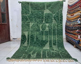 Könnte beinhalten: Ein großer, grüner Teppich mit geometrischem Muster und zotteliger Textur. Der Teppich hat ein Design aus Quadraten, Rechtecken und Rauten in verschiedenen Grüntönen. Der Teppich hat einen Fransenbesatz am unteren Rand und wird in Innenräumen ausgestellt.