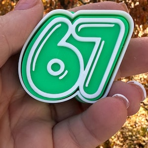 May include: A green and white number 67, with a white outline, is held in a hand. The number is a solid green color with a white border. The background is blurred, with fall foliage.