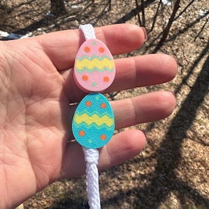 May include: Easter egg-shaped ornaments in pink and teal, decorated with zig-zag and dot patterns in yellow and orange. The eggs are connected by a white cord, perfect for Easter decorations.
