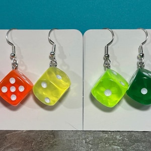 May include: A set of six pairs of dangle earrings featuring colorful dice charms. The dice are translucent and have white dots. The colors of the dice are pink, red, orange, yellow, green, and purple.
