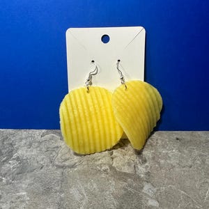May include: A pair of yellow chip-shaped earrings with silver hooks. The earrings are hanging from a white card.