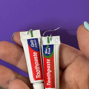 May include: A pair of silver earrings shaped like miniature toothpaste tubes. The tubes are red and white with green accents and the text "Toothpaste" and "Cavity protection" on one tube and "Toothpaste" and "Mint Fresh" on the other.