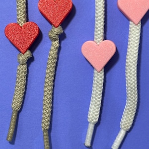 May include: Close-up of four shoelaces with heart-shaped charms. Two laces are tan with red hearts, and two are white with pink hearts. The background is a solid blue.