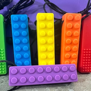 May include: Six colorful silicone teething toys shaped like Lego bricks. The toys are on a purple surface and have a black cord attached to them.