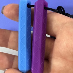 May include: Two sensory chew necklaces, one light blue and one purple, held in a hand. Each necklace is a long, hexagonal prism with a wavy pattern and a hole for a cord. The background is a solid light purple.