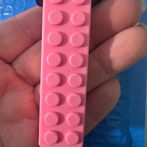 May include: A rectangular pink pendant with a textured surface featuring two parallel rows of eight raised circular studs, resembling a building block toy. The pendant is attached to a thin black cord, creating a simple necklace design.