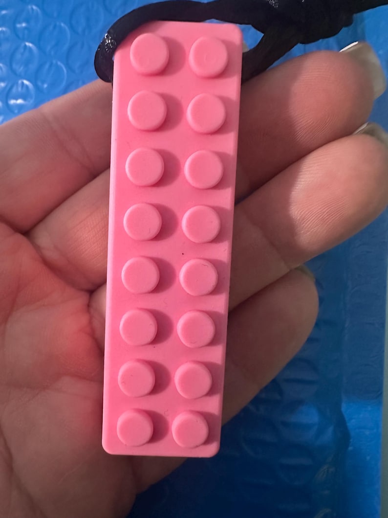 May include: A rectangular pink pendant with a textured surface featuring two parallel rows of eight raised circular studs, resembling a building block toy. The pendant is attached to a thin black cord, creating a simple necklace design.