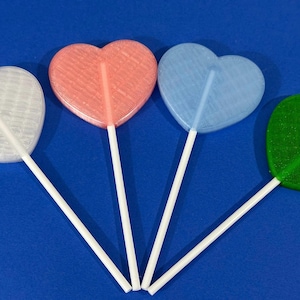 May include: Four heart-shaped lollipops in white, coral, light blue, and green, each with a white stick. The lollipops are arranged against a solid blue background, creating a colorful and playful image.