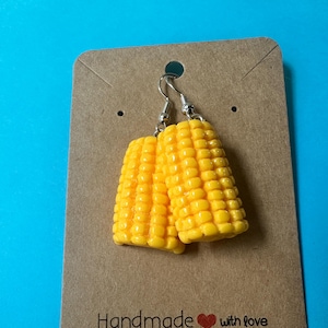 May include: A pair of novelty earrings shaped like yellow corn on the cob, hanging from silver-colored hooks. The earrings are displayed on a brown card with the text "Handmade with love" at the bottom, against a bright blue background.