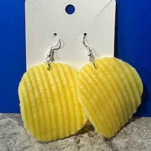 May include: A pair of novelty earrings featuring a yellow, ridged potato chip design. The earrings hang from silver-colored fish hook ear wires and are attached to a white card with a blue background.