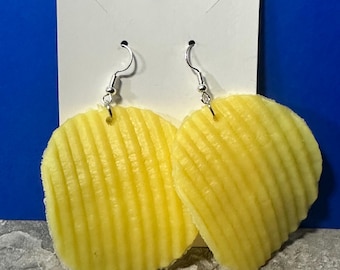 May include: A pair of novelty earrings featuring a yellow, ridged potato chip design. The earrings hang from silver-colored fish hook ear wires and are attached to a white card with a blue background.