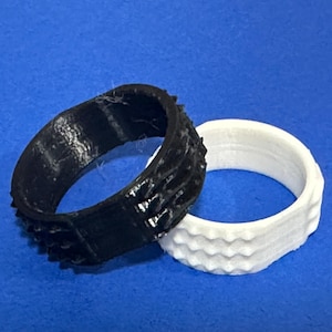 May include: Two rings, one black and one white, rest on a blue surface. The black ring has a textured, spiky design. The white ring has a wavy, textured pattern. Both rings appear to be made of a solid material.