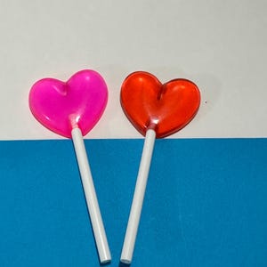 May include: Two heart-shaped lollipops on white and blue backgrounds. The lollipop on the left is pink and the lollipop on the right is red.
