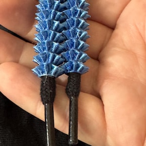 May include: Close-up of two blue decorative drawstring ends. Each end features a series of layered, fan-like shapes in a vibrant blue hue. The cord ends are black and attached to a black cord.