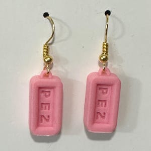May include: A pair of pink Pez dispenser earrings. Each earring features a rectangular pink charm with the word "PEZ" embossed in white. The earrings have gold-colored ear wires and small gold beads.
