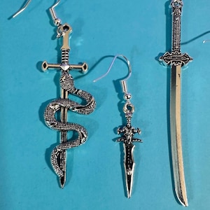 May include: Three pairs of silver-colored earrings with different sword designs. One earring features a snake wrapped around a sword, another a dagger, and the third a long sword. The earrings are displayed on a blue background.