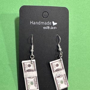 May include: Silver-tone dangle earrings featuring miniature stacks of $100 bills. The earrings are attached to a black card with the text "Handmade with love". The background is a solid green color.